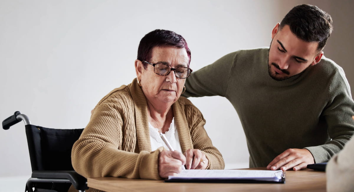 An elderly woman in a wheelchair signing documents with a younger man leaning over her
