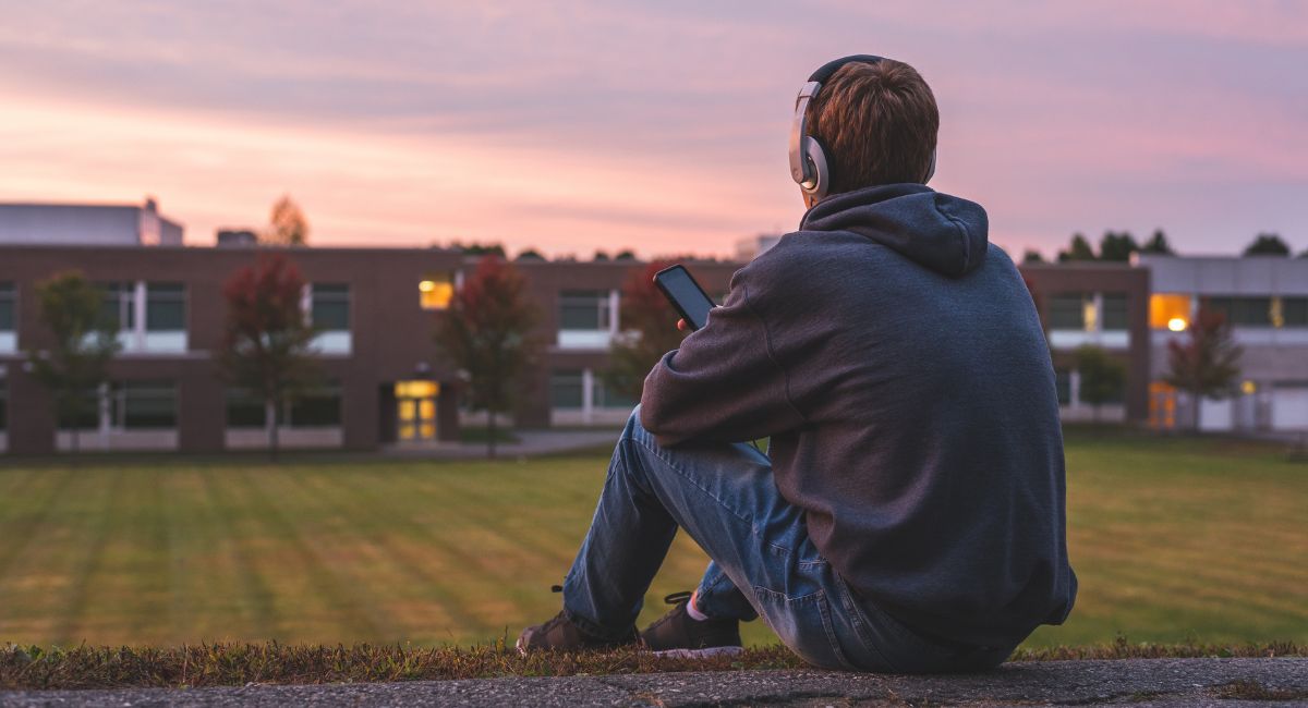 A young man with headphones sitting on a field alone as the sun sets