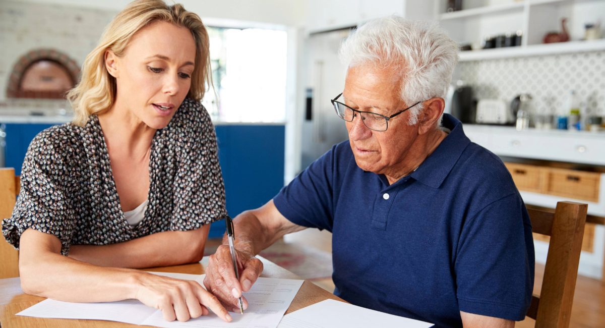 An elderly man signing some documents with a younger woman pointing at the document