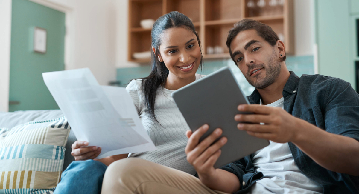 A couple sitting on a couch looking at a tablet, holding documents