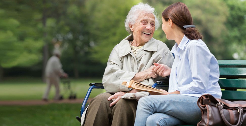 Older woman, sitting on park bench, laughing with care worker 