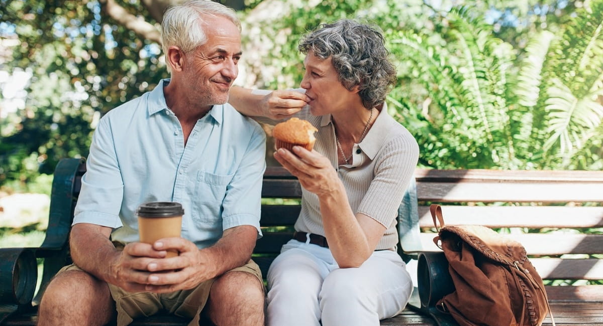 Older man and woman enjoying coffee and a muffin on park bench.