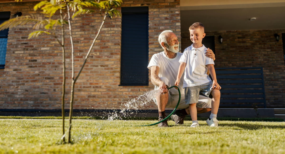 An older man with his grandson watering a tree in the backyard
